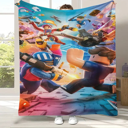 A person holds up a large fleece blanket featuring a colorful, dynamic cartoon battle scene with block-style characters, vehicles, and bright action effects. The blanket is shown in a home setting with a window and floor visible in the background.