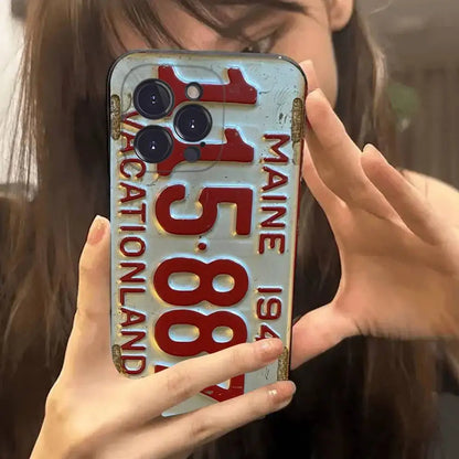 A close-up of a person holding a phone case designed to look like a vintage Maine vacation land license plate with red numbers and text on a distressed light blue background.