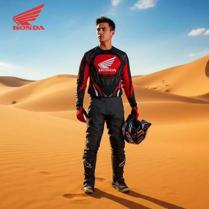 A man stands in a desert dune wearing a red, black, and white Honda motocross jersey and pants, holding a helmet under one arm. The Honda logo is visible in the top left corner.