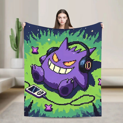 A woman holds up a large blanket featuring a pixel-art purple creature wearing headphones on a green grassy background, photographed in a modern indoor room with a cactus and sofa visible.