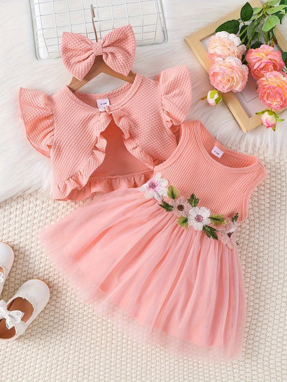 Pink children's dress with floral details on a textured surface with flowers and a bow in the background.