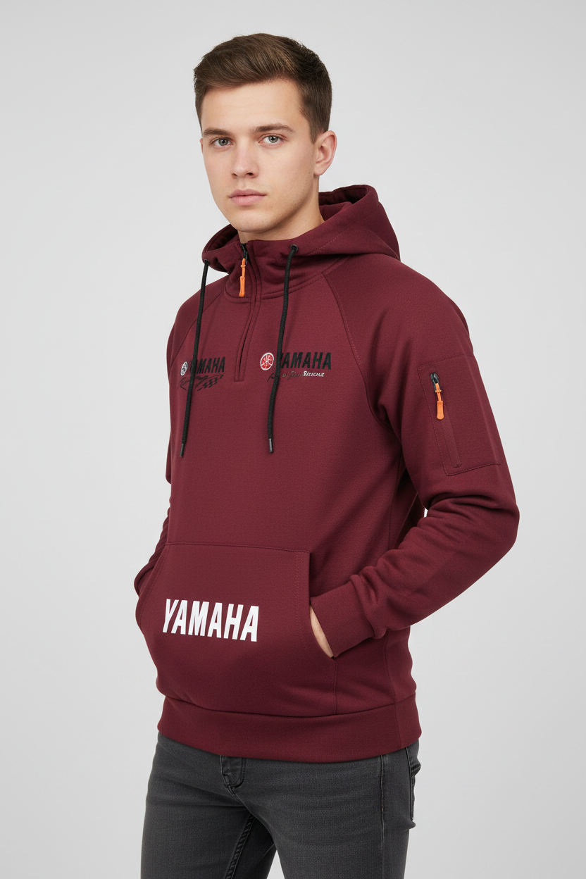 A man wearing a maroon Yamaha pullover hoodie with black drawstrings, black and white logo graphics on the chest, and a front kangaroo pocket, paired with dark gray jeans against a light gray studio background.