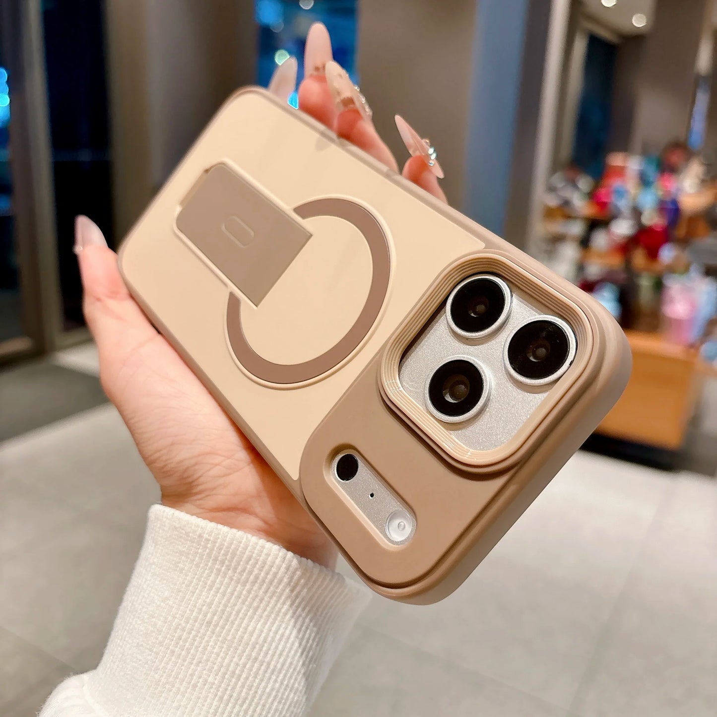 A hand holding a beige phone case with a built-in kickstand and MagSafe ring, photographed in a blurred indoor retail environment.