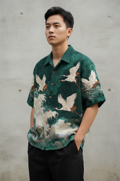 A man wearing a dark green short-sleeve button-up shirt with a detailed crane and wave pattern, paired with black pants, standing against a textured gray concrete wall.