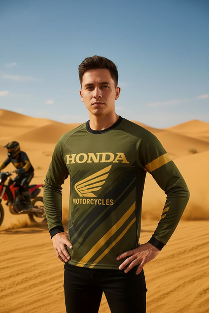 A man wearing a green and gold long-sleeve Honda Motorcycles jersey stands in a desert setting with sand dunes and a motorcycle rider in the background.