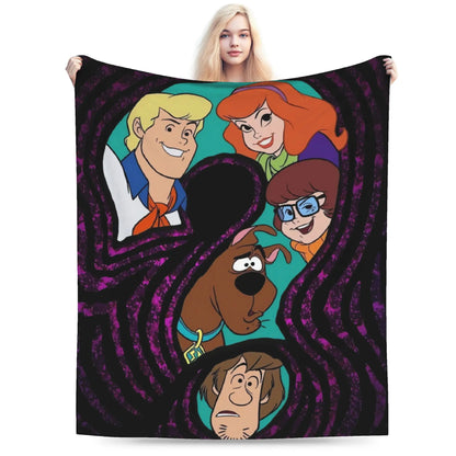 A person holds up a large square blanket or throw featuring cartoon characters from Scooby-Doo. The blanket has a black and purple swirling maze-like pattern with character portraits of Fred, Velma, Shag, and Scooby-Doo on a teal background.